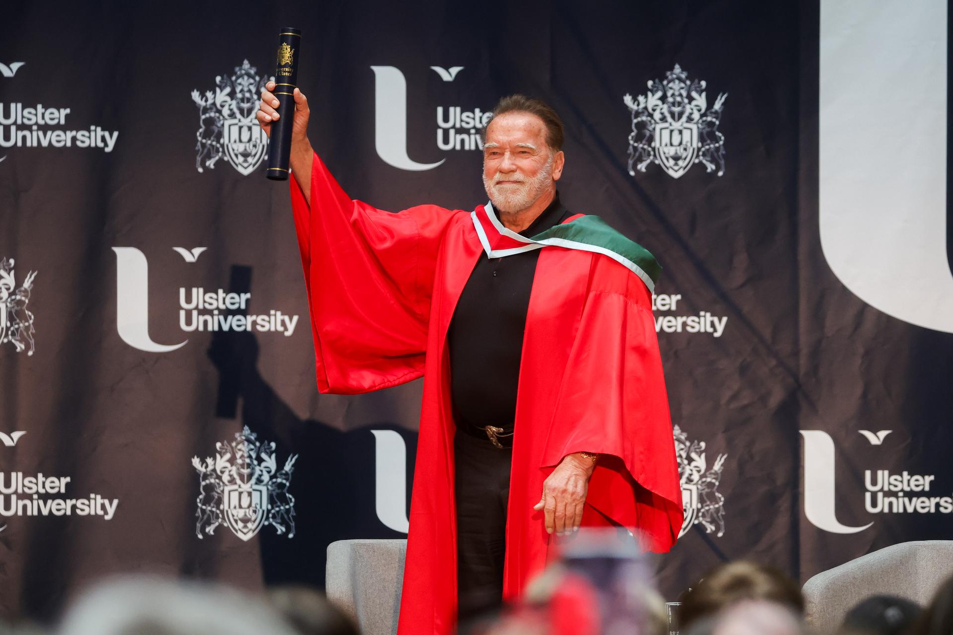 A person in a red graduation gown holds up a diploma, smiling, in front of an 